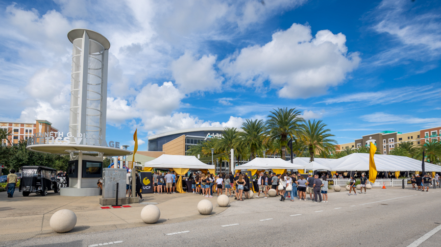 Outdoor campus event with white tents, people lined up and palm trees under a bright blue sky.