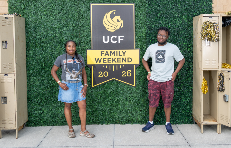 Two people stand beside a UCF Family Weekend 2026 sign against a green hedge backdrop with lockers on either side.