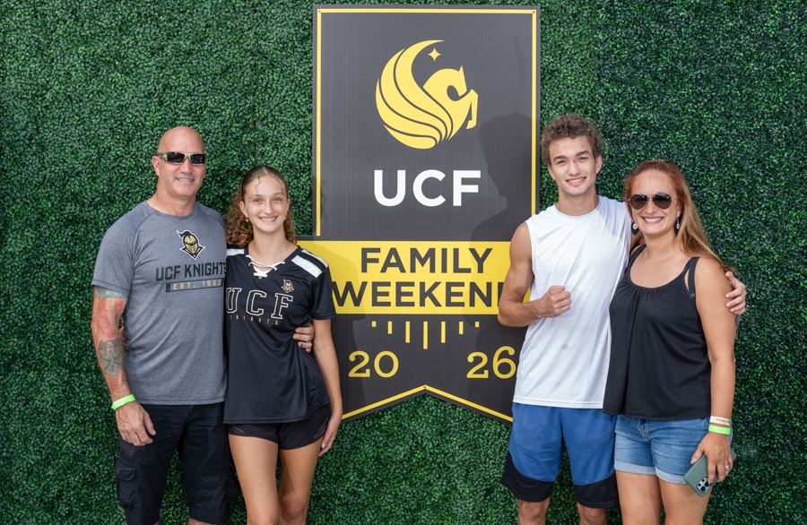 Family of four posing in front of a UCF Family Weekend banner on a green hedge backdrop (2026).