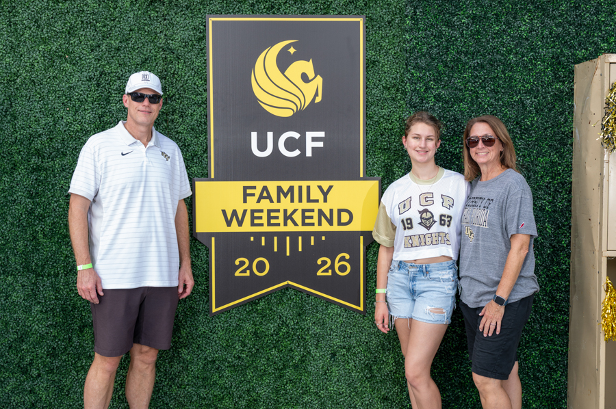 Three people pose beside a large UCF Family Weekend sign on a green hedge backdrop: a man in a white polo and cap on the left, a young woman in a Knights shirt in the middle, and a woman in sunglasses on the right.
