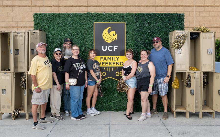 Eight people wearing UCF gear posing in front of a UCF Family Weekend sign, with lockers on both sides and a green hedge backdrop.