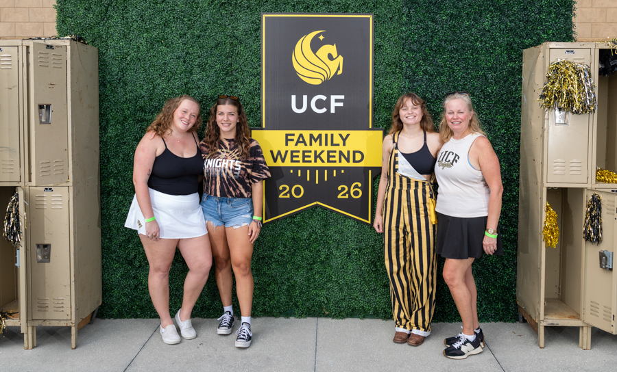 Four women pose and smile in front of a UCF Family Weekend 2026 banner, with lockers and a green hedge backdrop nearby.