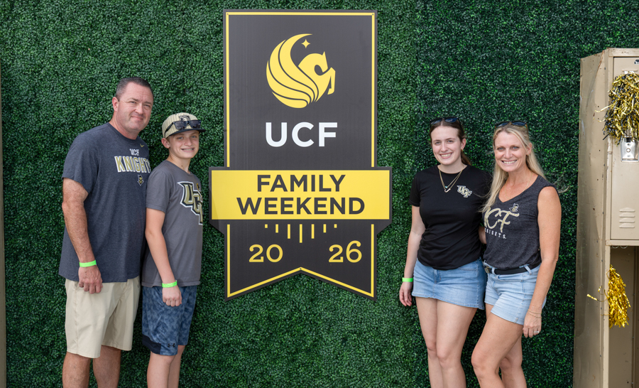 Family of four posing beside a UCF Family Weekend sign on a green hedge backdrop (2026).