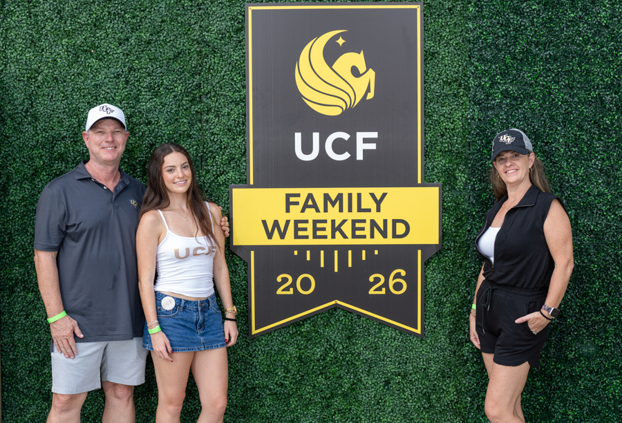 Three people pose beside a tall UCF Family Weekend 2026 sign against a green hedge backdrop.