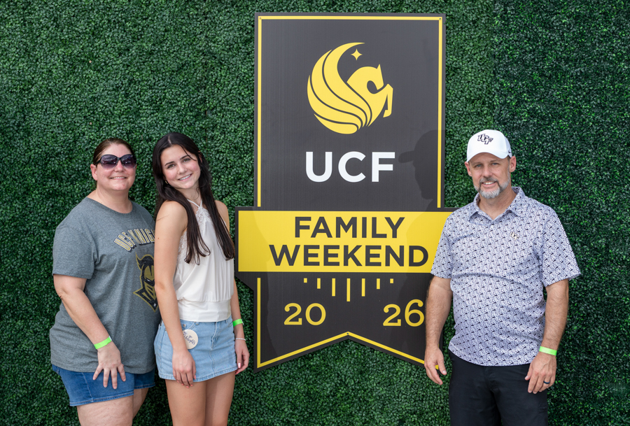 Three people pose beside a large UCF Family Weekend sign on a green hedge backdrop, 2026 event.