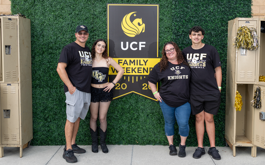 Four people wearing UCF Knights apparel stand together in front of a large UCF Family Weekend banner against a green hedge background.