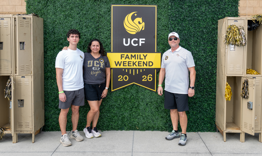 Three people pose in front of a UCF Family Weekend 2026 banner on a green hedge backdrop, with beige lockers on both sides (a young man left, a woman center, and a man right).