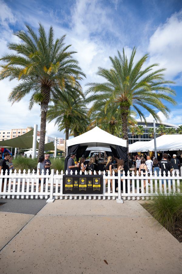 Outdoor market or festival scene under palm trees with white canopy tents and a white picket fence, people mingling around stalls.