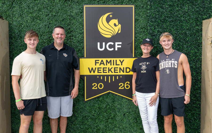 Four people pose in front of a UCF Family Weekend 2026 sign against a green hedge backdrop. Left to right: two teens and two adults, smiling.