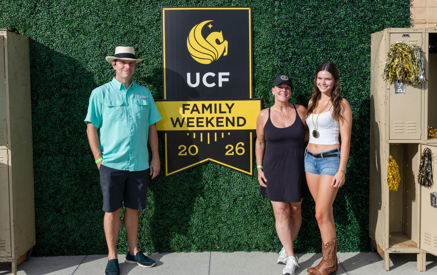 Three people pose in front of a UCF Family Weekend 2026 sign against a green hedge backdrop, smiling outdoors.