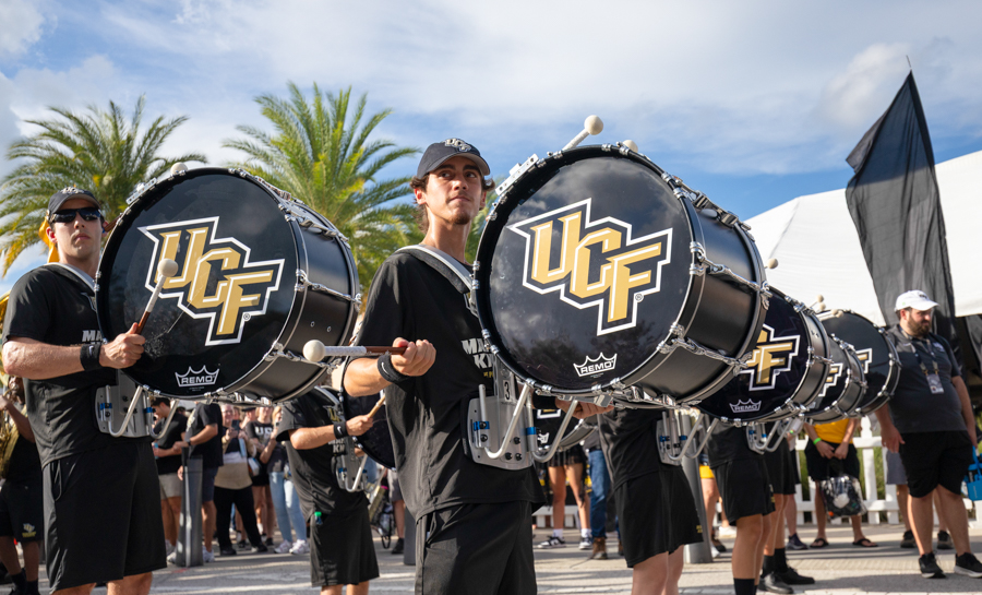 Marching band drummer holding a large bass drum with a UCF logo during an outdoor parade; palm trees in the background.