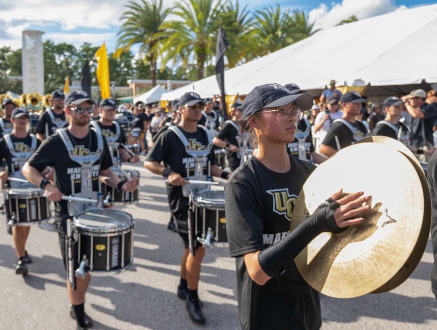 Drumline performers marching outdoors at an event, foreground musician holding a large cymbal in a black-uniforms band.