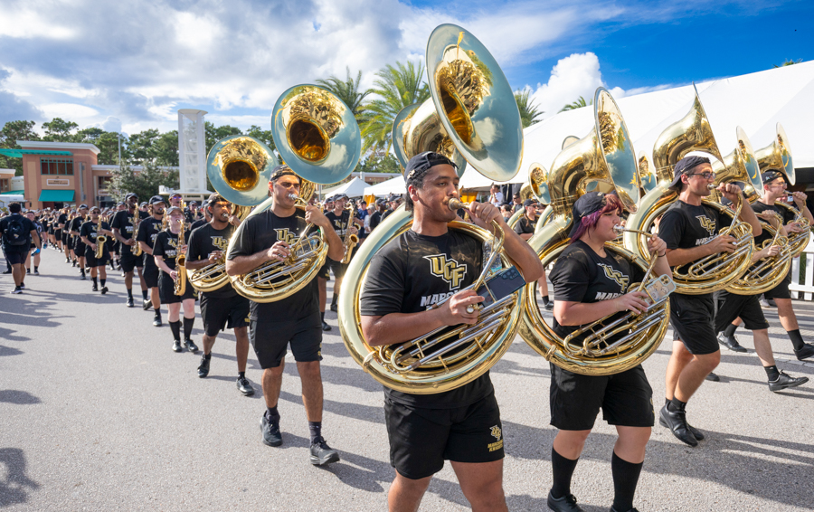 University of Central Florida marching band members march with sousaphones and brass instruments in a sunny outdoor parade outdoors.