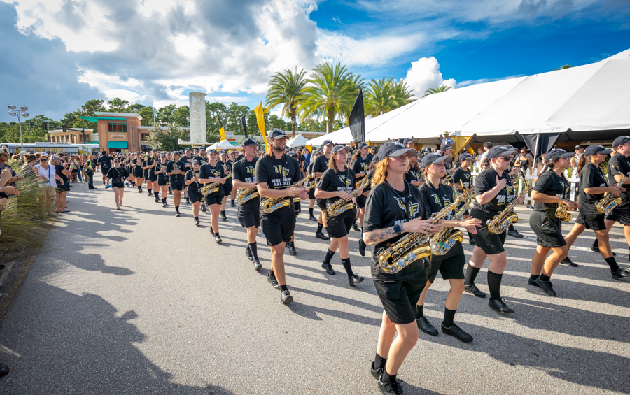 Marching band in black uniforms marches with saxophones at an outdoor parade, with tents and palm trees in the background.