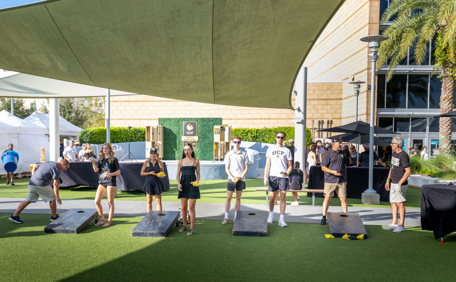 Group of young people lined up at cornhole boards at an outdoor campus event under a shade sail, with a UCF backdrop in the background.