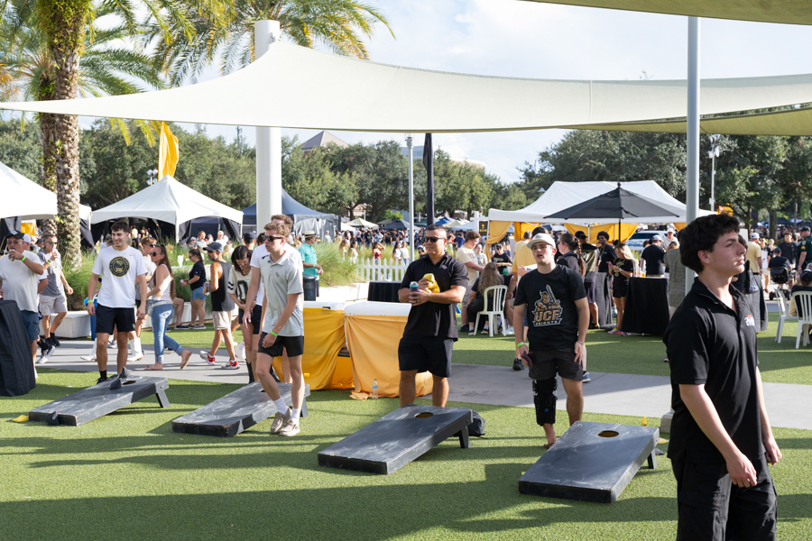 People participate in an outdoor festival game area, tossing bags onto slanted boards under shade sails and tents.