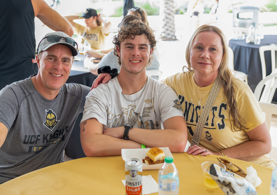 Three friends sit at a yellow table outdoors, smiling, wearing UCF Knights shirts with a burger and drinks in front of them.
