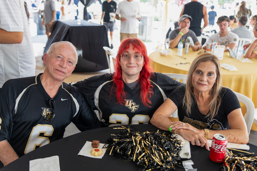 Three people in UCF jerseys sit at a round table with snacks and black-gold decorations at a campus event.