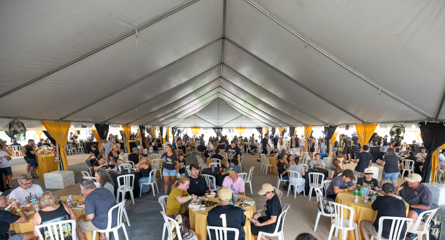 Crowded banquet under a large white event tent with dozens of round tables and guests dining.