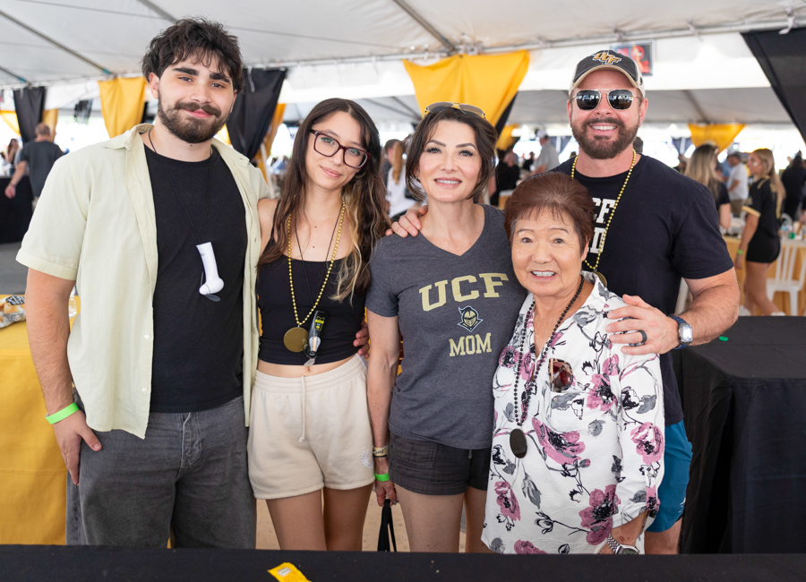 Group of five smiling people posing together at an outdoor event under a yellow-and-black tent. First man on left wears a light shirt, second woman with glasses, center woman in UCLA Mom shirt, older woman in floral blouse, and man on right wearing a cap and sunglasses.