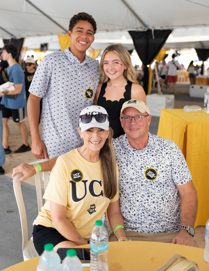 Four people posing together at an outdoor event under a white tent, smiling for the camera; two are seated at a yellow table wearing UC gear.