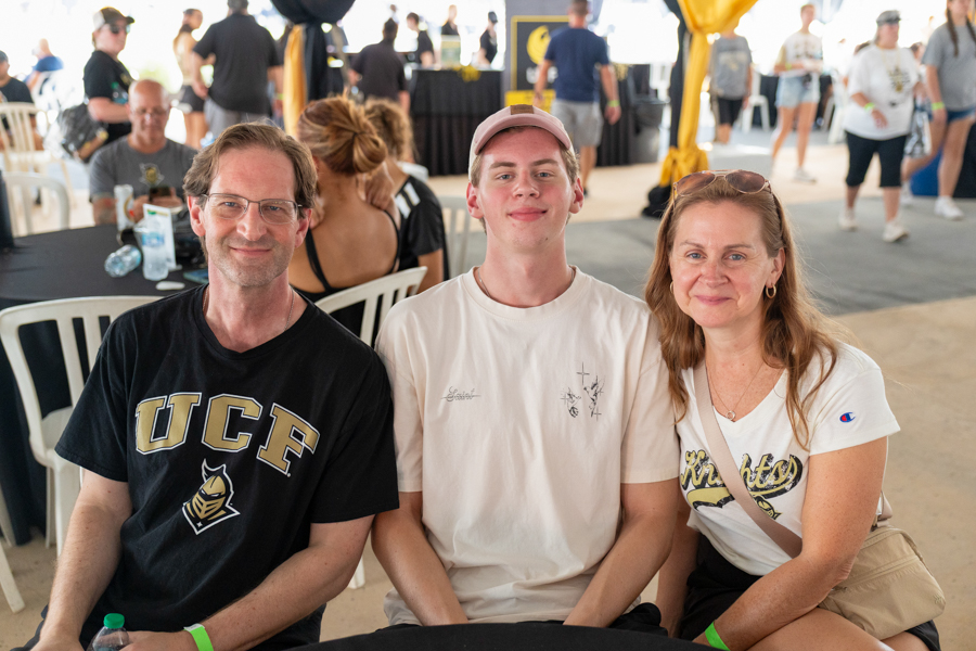 Three people sit together at a round table at a busy indoor event, smiling for the camera; left man in a black UCF shirt, center young man in a white tee and pink cap, right woman in a white shirt with sunglasses on her head.