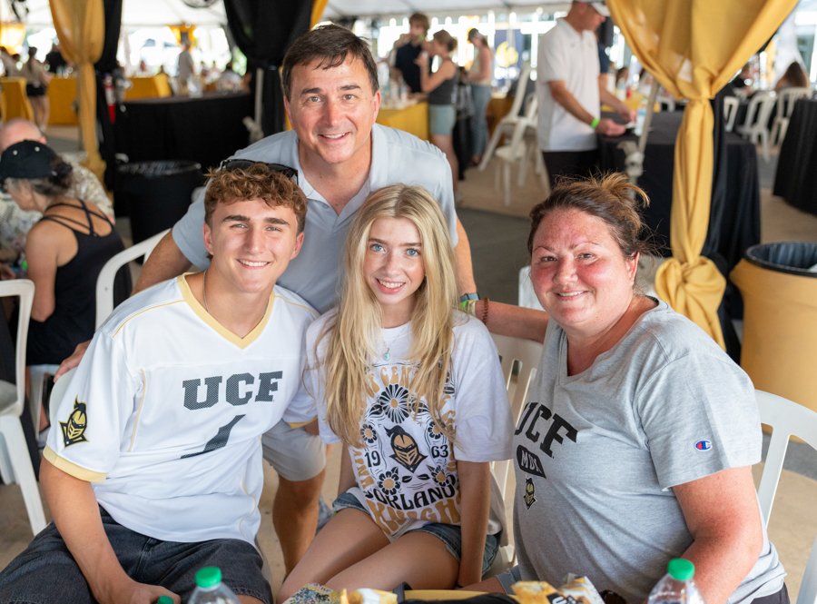 Four people pose for a group photo at an outdoor event under yellow canopies; two wear UCF shirts and all are smiling.