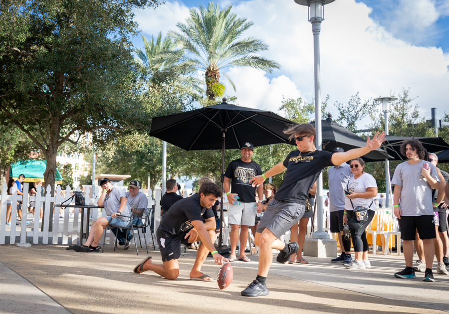 Two young men playfully punt a football while a crowd watches under umbrellas at a sunny outdoor event.