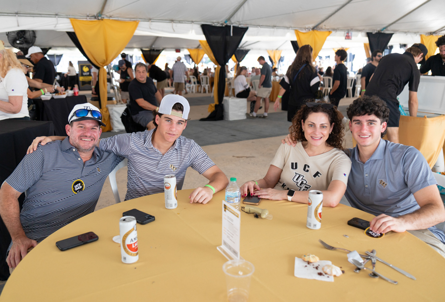 Four friends sit around a yellow-tablecloth table under a tent at an outdoor event, smiling with drinks and phones in hand.