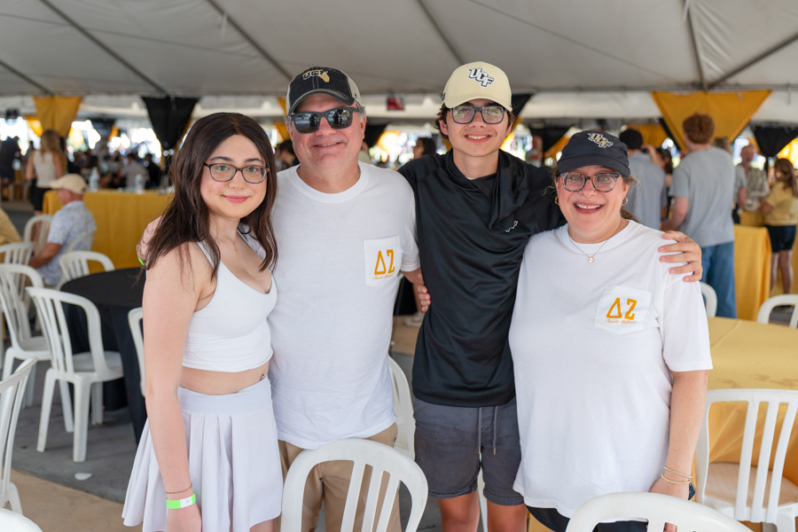 Four people pose for a photo at an outdoor event under a tent; two wear white shirts with ΔΖ logos and all smile at the camera.