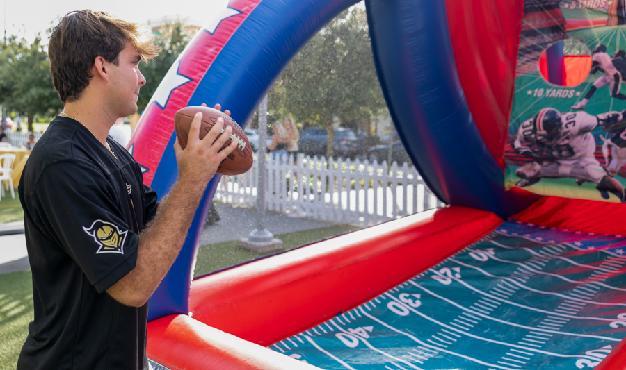 Young man in a black shirt holding a football inside an inflatable football-themed game at an outdoor event on a sunny day.
