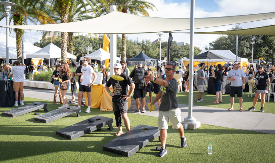 Man tossing a bean bag toward a cornhole board at an outdoor festival; tents, palm trees, and attendees in the background.