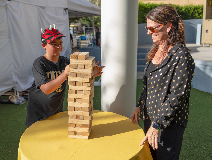Boy wearing a horned costume builds a wooden Jenga tower on a yellow table as a woman smiles nearby outdoors.