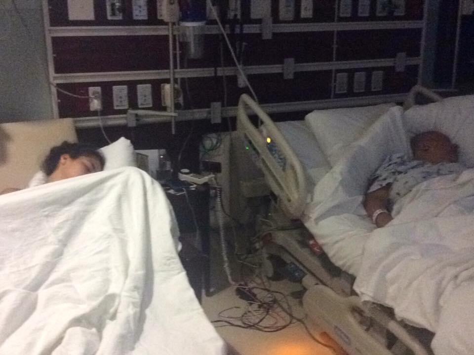 Two patients rest in hospital beds in a dimly lit room, with monitors and cables nearby.