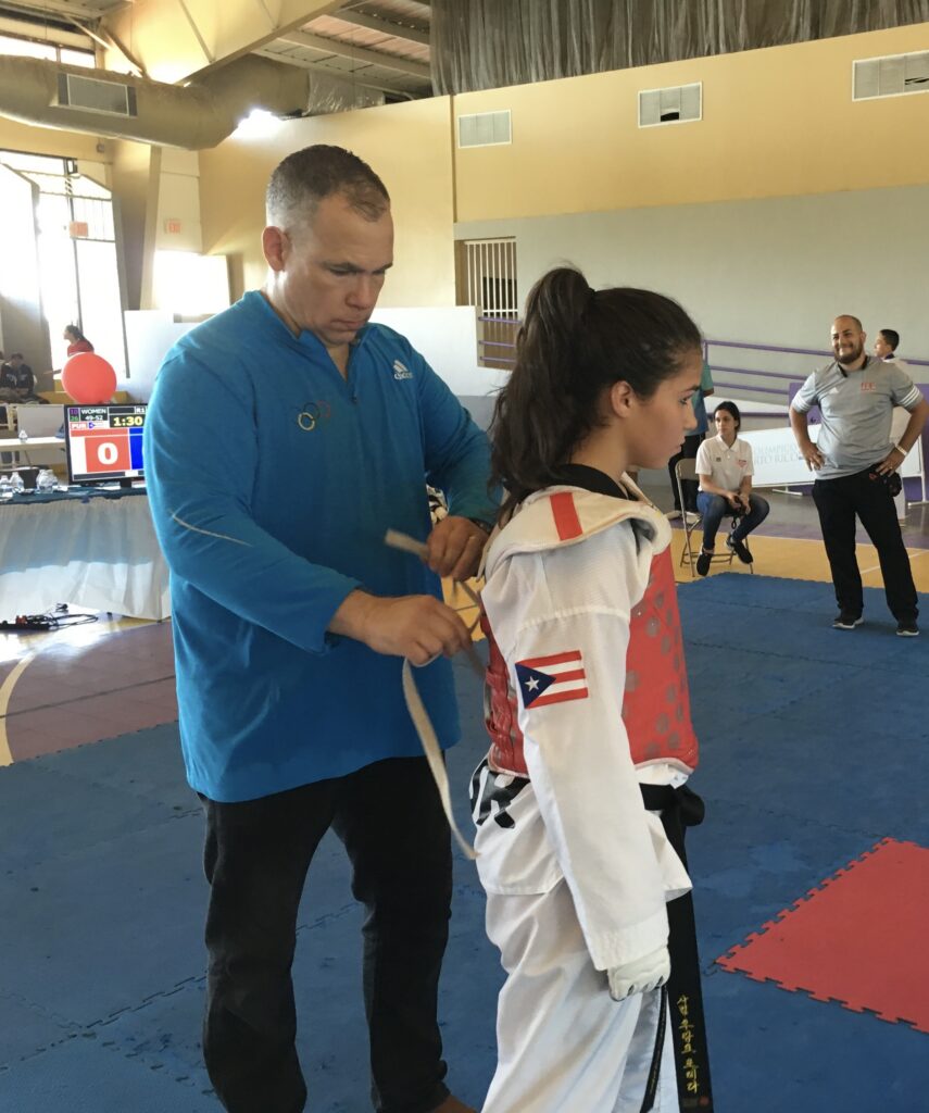 Coach in a blue jacket ties a young taekwondo competitor's gear, a red chest guard visible, in a gym.