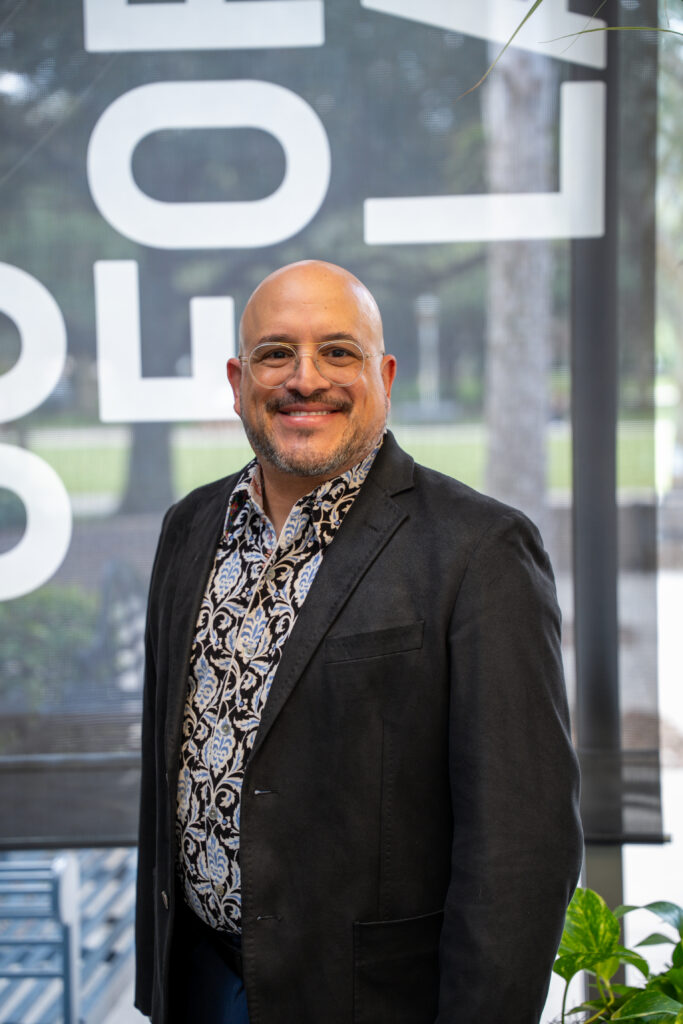 Smiling bald man wearing round glasses, a patterned shirt, and a black blazer indoors with large window panels behind him.