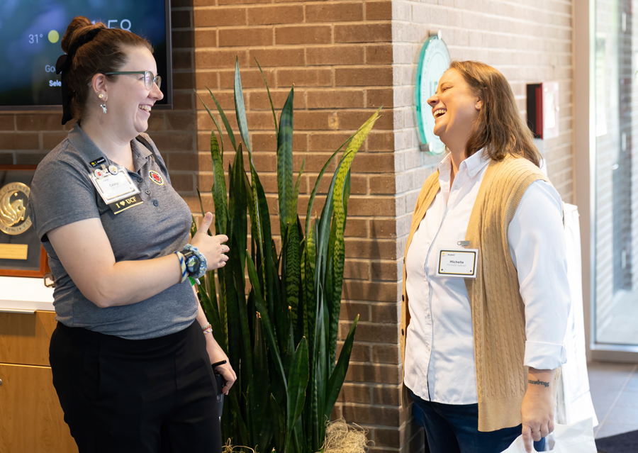 Two coworkers laugh and chat near a tall snake plant in a brick-walled lobby, both wearing name badges.