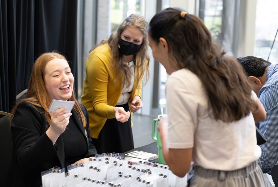 A red-haired woman laughs while handing a name badge to a visitor at a registration table; another staff member wearing a mask assists nearby. May be an event check-in scene.