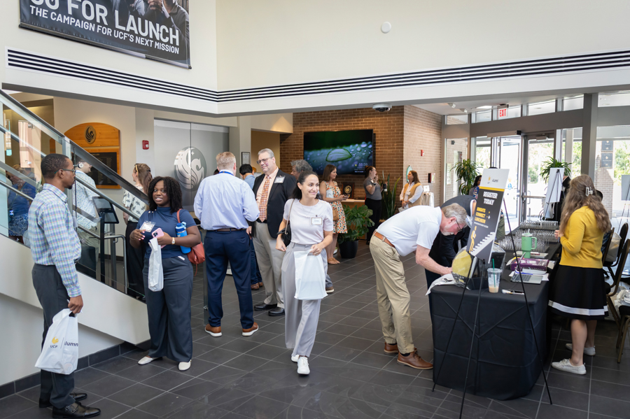 Diverse professionals mingle in a modern lobby during a launch event; a registration table with staff is on the right and attendees hold tote bags.