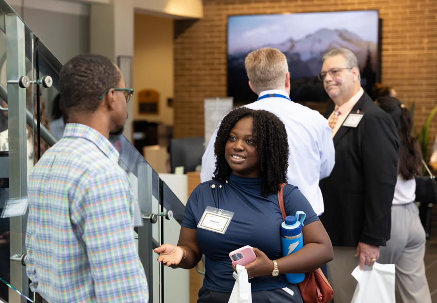 Women networking at a conference, speaking with a man by a glass railing, name badge visible