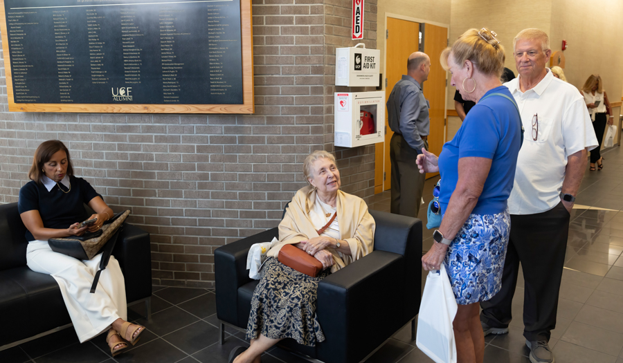 Elderly woman in a beige shawl sits in a black chair in a brick lobby, talking with a standing woman in blue; a man and another seated woman are nearby, with an alumni board on the wall.