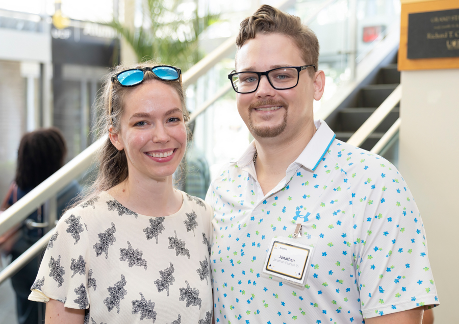 Two smiling attendees posing for a photo at an indoor event; woman wears a patterned top with blue sunglasses on her head, man wears glasses and a floral shirt with a name badge reading Jonathan nearby.