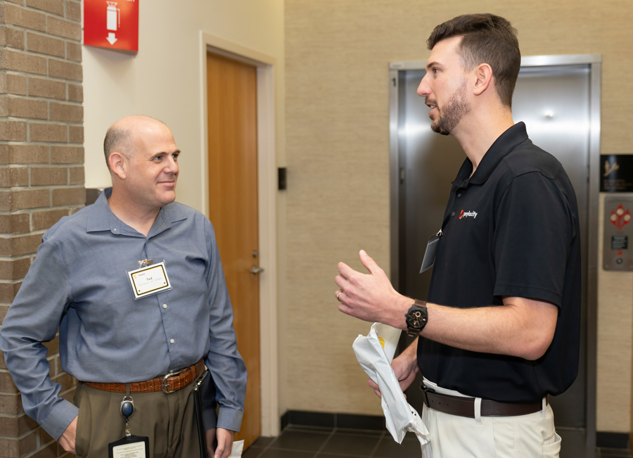 Two men in an office hallway conversing; one in a blue shirt with a name badge, the other in a black polo holding papers and gesturing.