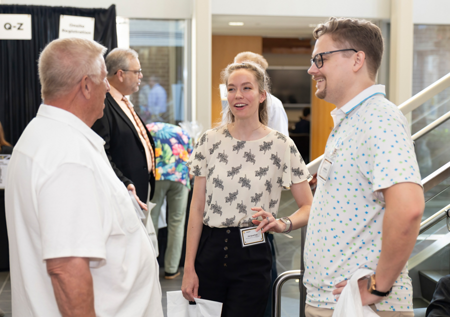 People conversing at a conference or expo, a young woman in the center with a name badge talking to two men.