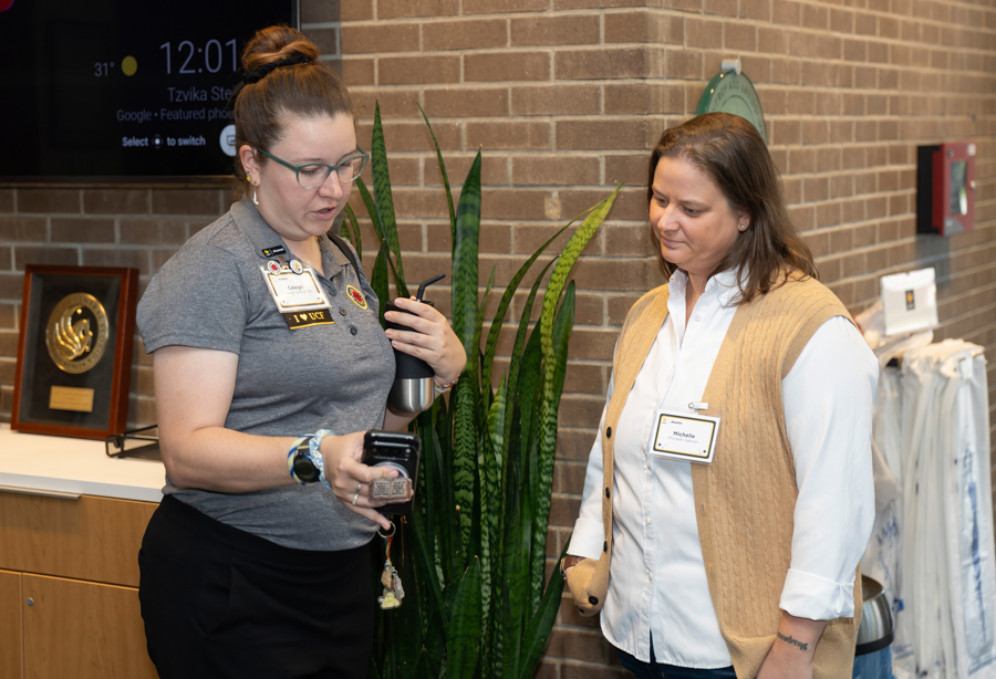 Two women in a hallway, one showing a phone to the other near a tall plant and brick wall