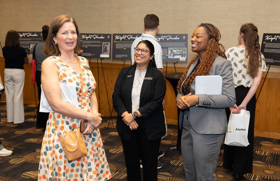 Three women in business attire talk and smile at a conference, with informational posters behind them on display boards that read Knight Chadus (sic) or similar.