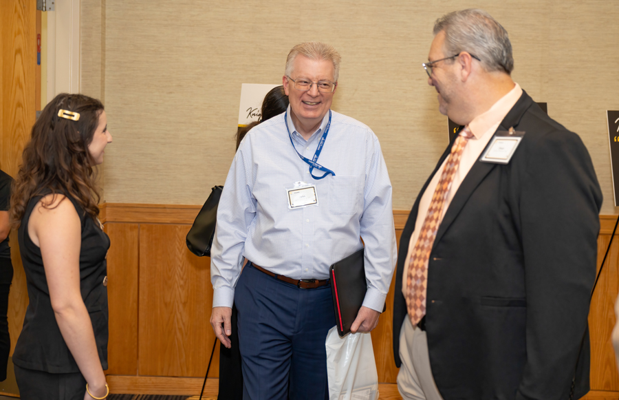 Three professionals chatting at a conference; man in a light blue shirt smiles, wearing a name badge and holding a folder and bag.