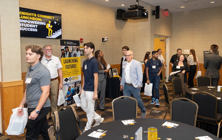 People with name badges walk through a college career expo, passing posters that read 'Launching Futures' and 'Knights Connect Luncheon'.