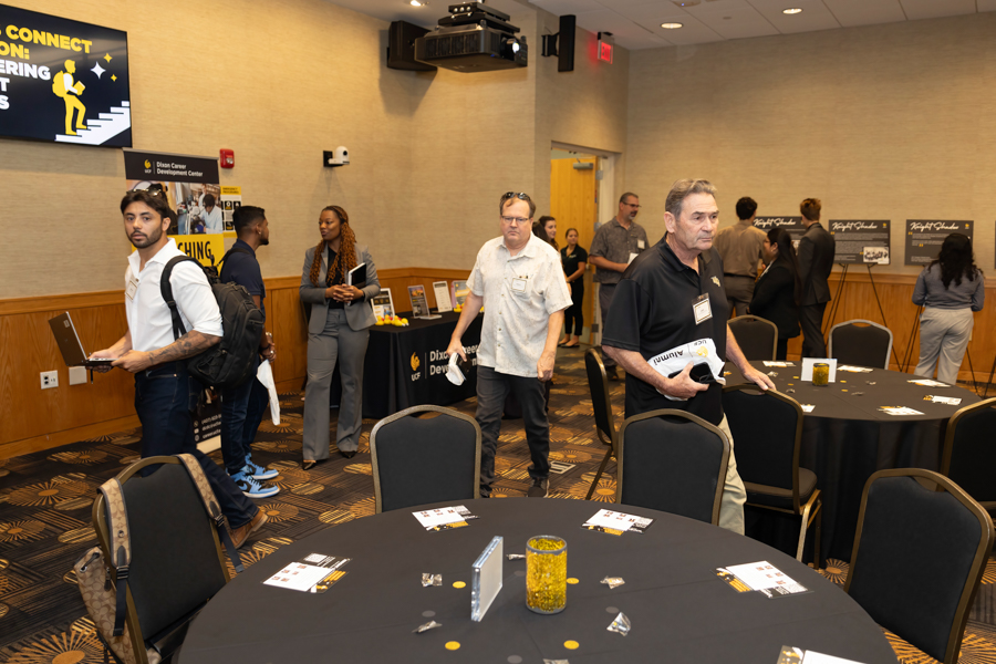 Professionals mingle at a conference expo with round tables, booths, and banners in a hotel ballroom.