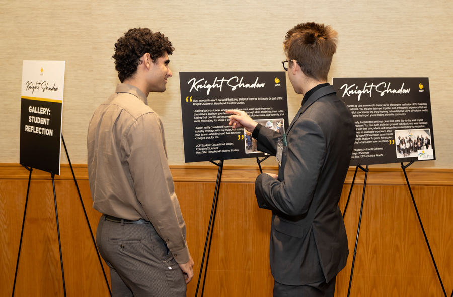 Two people in business attire discuss display boards at a gallery, with Knight Shadow posters on stands behind them.